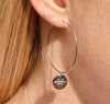 Right ear of a person wearing a large, thin silver hoop earring with round embossed pendant with star detail