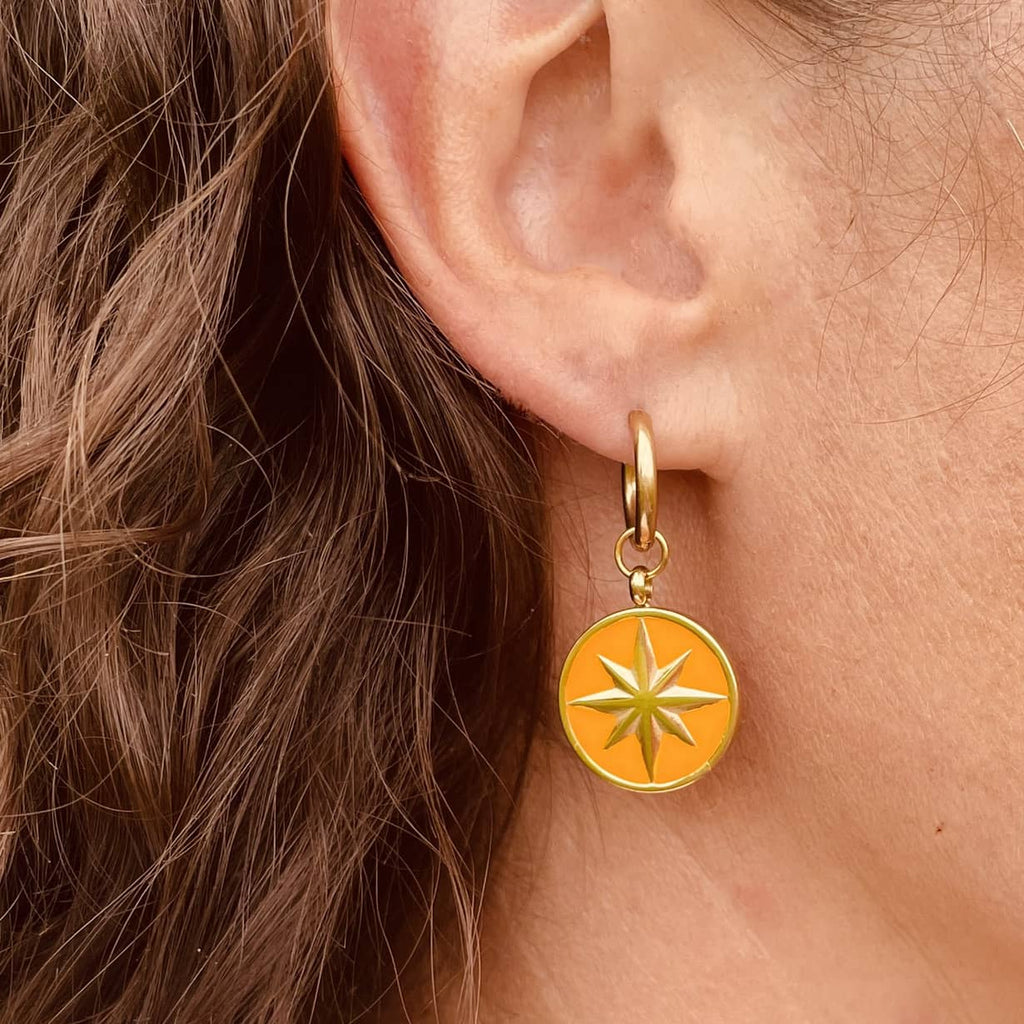 Right ear of a person wearing a small golden huggie hoop earring with a round pendant in golden yellow and a gold star
