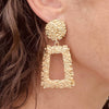 Right ear of a person wearing a statement earring in textured gold effect with a large round stud and rectangular pendant