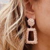 Right ear of a person wearing a statement earring in textured rose gold effect with a large round stud and rectangular pendant