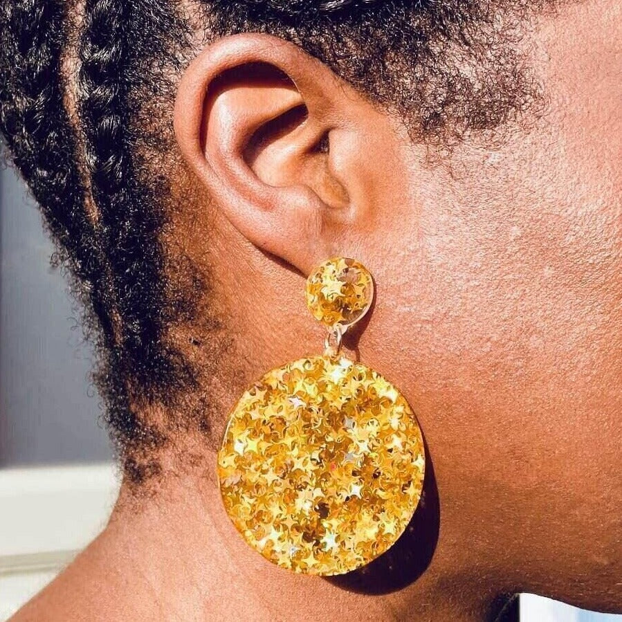 Right ear of a person wearing a large, acrylic statement pierced earring with circular stud and round disc drop pendant and golden yellow metallic star detail