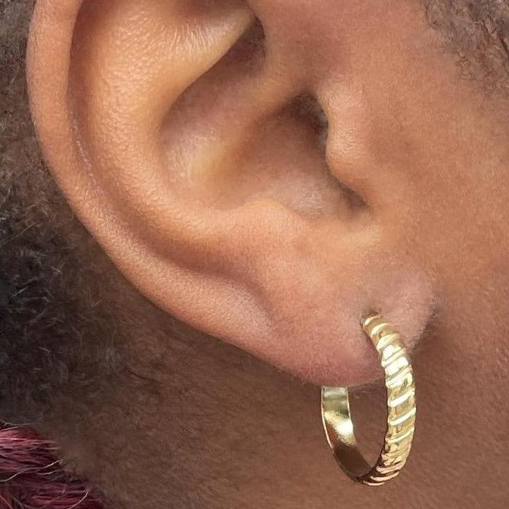 Right ear of person wearing small gold hoop pierced earring with striped embossed detail