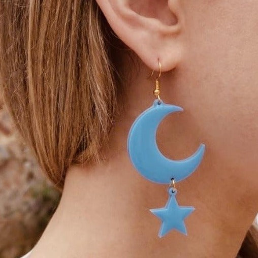 Right ear of a person wearing a pale blue resin statement pierced earring with gold hook clasp and moon and star shaped pendant design