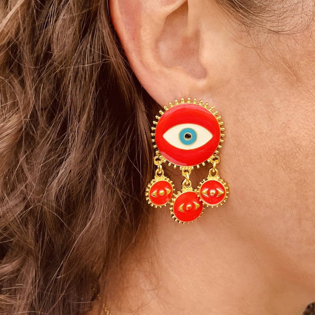 Right ear of a person wearing a large red enamel and gold statement dangle pierced earring with a large round disc with an evil eye design and three small red disc pendants